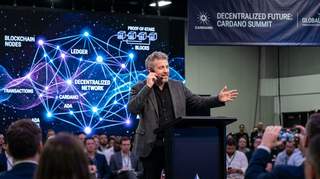 Charles Hoskinson speaking at conference with decentralized computing network visualization in background
